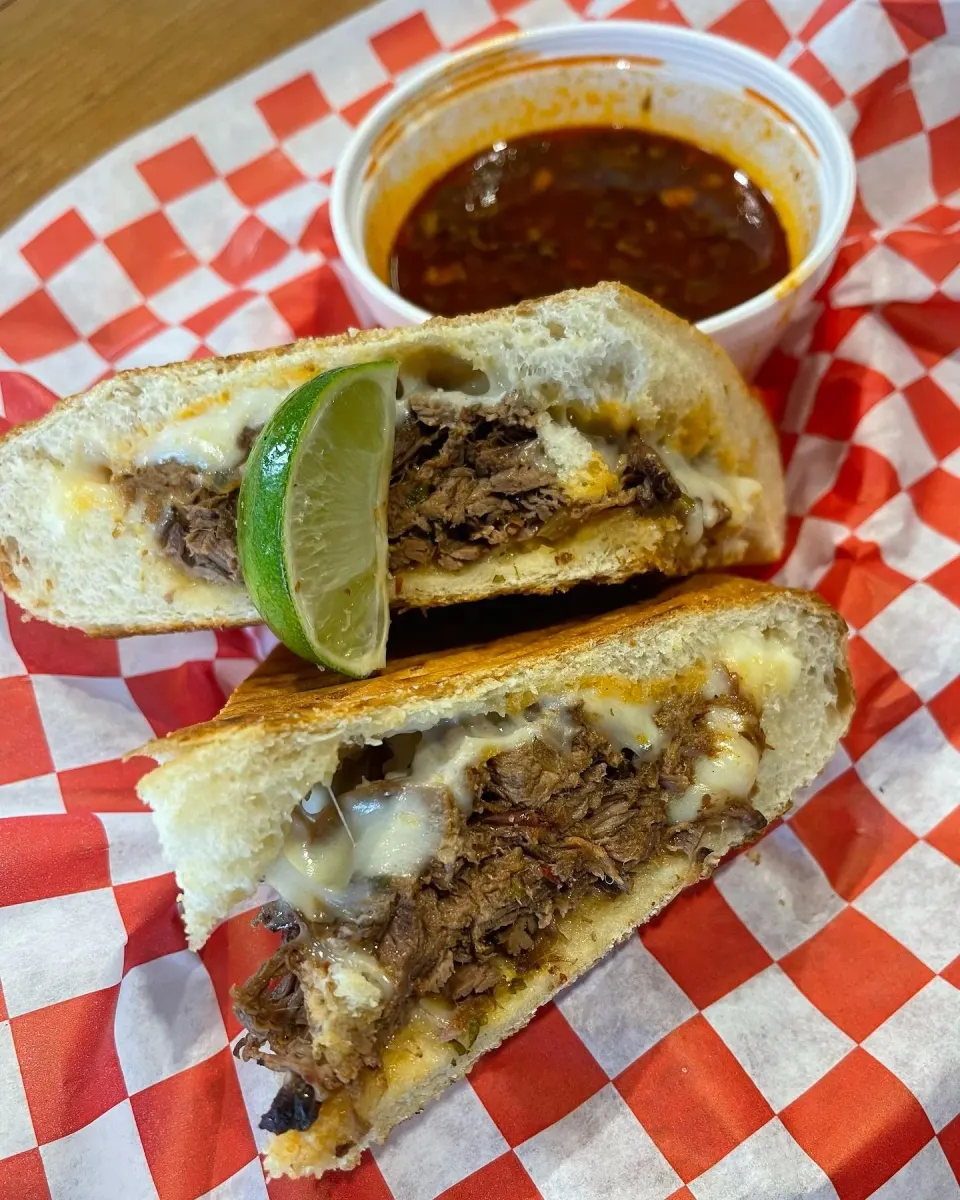 A sliced beef chimichanga with melted cheese and a lime wedge, accompanied by a side of red chili sauce on red and white checkered paper.