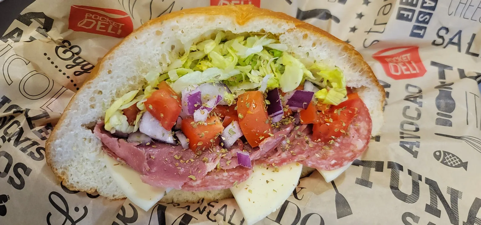Sub sandwich with lettuce, tomatoes, onions, ham, salami, and cheese inside a hoagie bun, on branded paper wrapping.