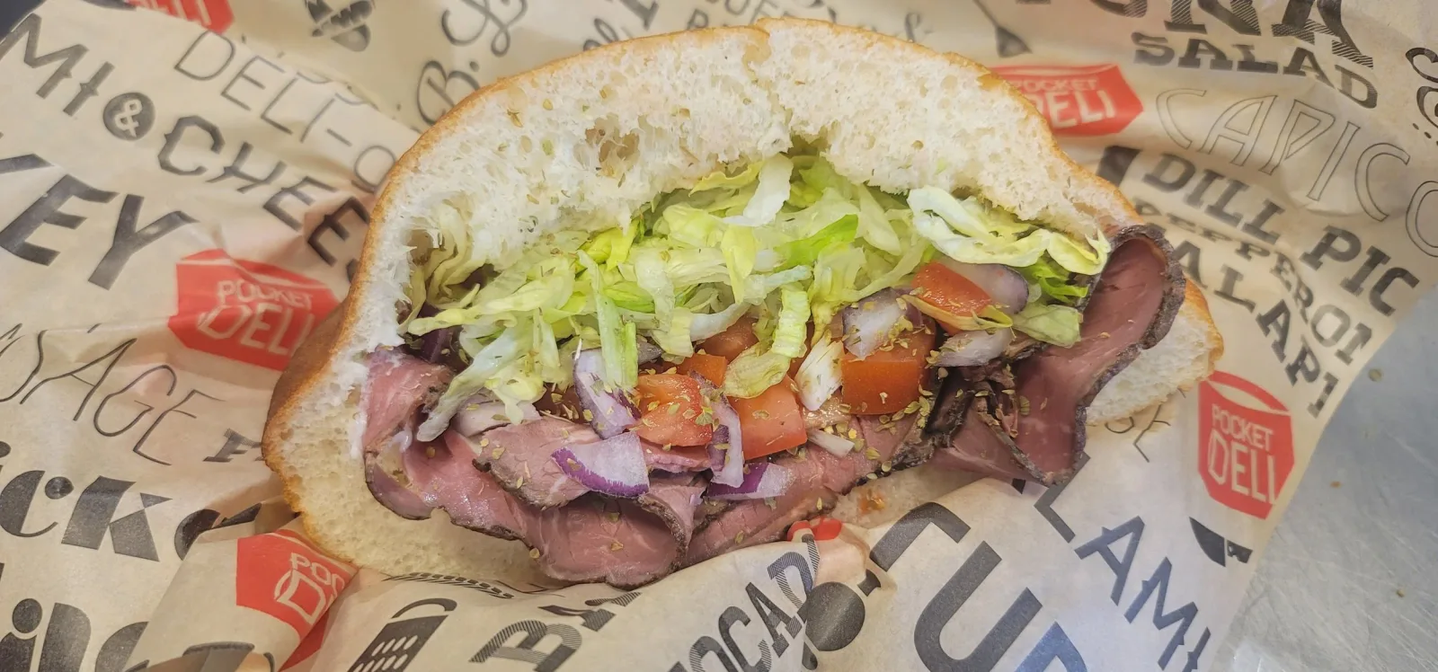 A sandwich with roast beef, chopped lettuce, tomato slices, red onion, and green herbs in a hoagie bun, wrapped in branded paper.